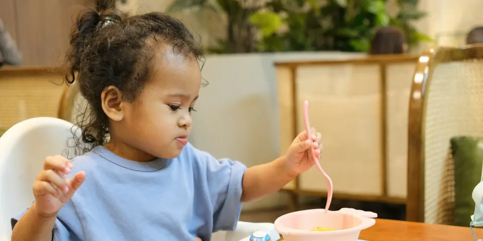 Niña pequeña en su silla de comer con una cuchara y un tazón
