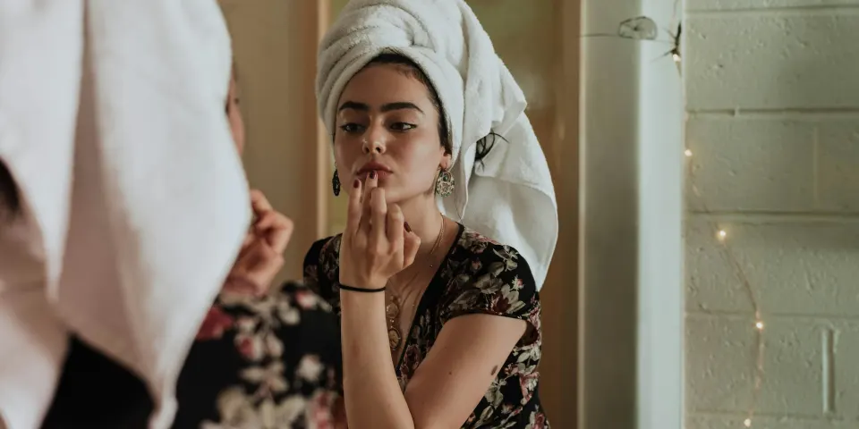 Mujer maquillándose frente al espejo con una toalla en el cabello