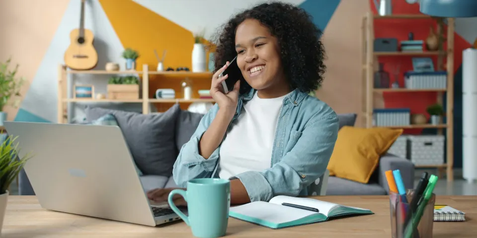 Mujer sonriente hablando por celular mientras trabaja desde su casa con el portátil